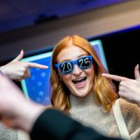 A grad smiles and points while wearing 2025 glasses